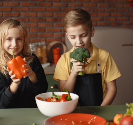 Cute girl and boy with fresh pepper and broccoli in hands standing near desk with assorted vegetables and salad bowl during vegetarian meal preparation