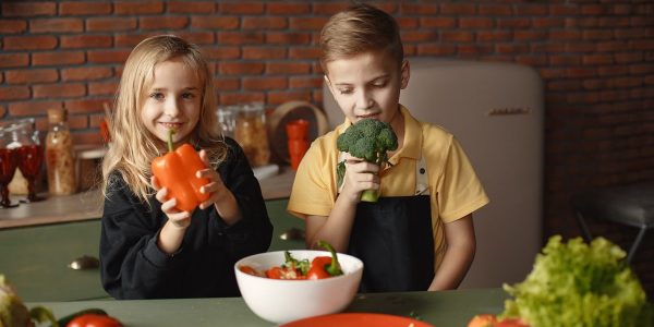 Cute girl and boy with fresh pepper and broccoli in hands standing near desk with assorted vegetables and salad bowl during vegetarian meal preparation